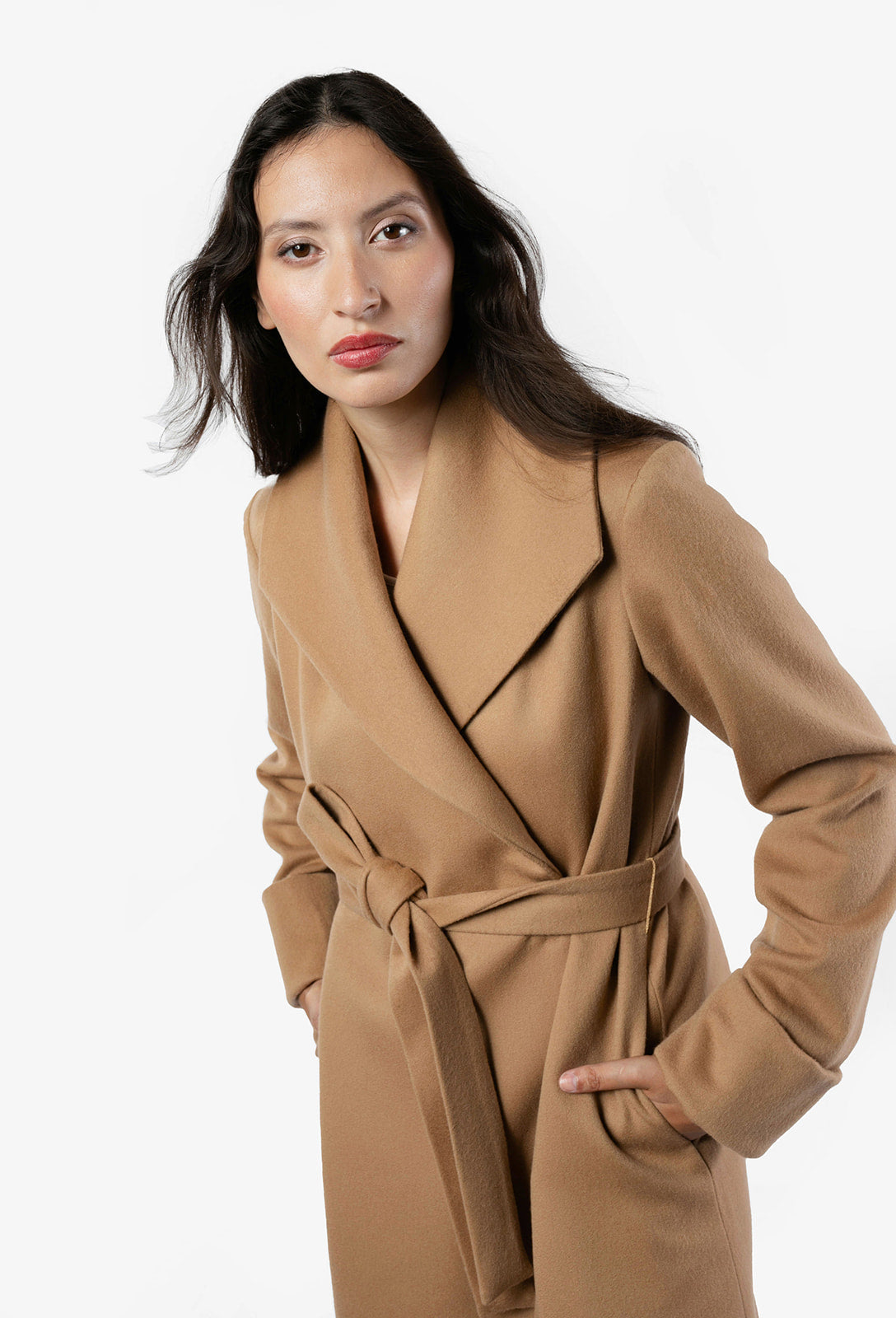 A close-up image of a woman in a camel coat. The woman is turned to the sided, showcasing its belted design and belt loop. The coat features cuffed long sleeves. The woman's hand is casually tucked into one of the coat's pockets, emphasizing its stylish and functional details. The coat features an oversized collar and is made from 100% wool fabric in a camel color.
