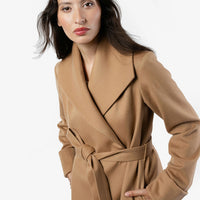 A close-up image of a woman in a camel coat. The woman is turned to the sided, showcasing its belted design and belt loop. The coat features cuffed long sleeves. The woman's hand is casually tucked into one of the coat's pockets, emphasizing its stylish and functional details. The coat features an oversized collar and is made from 100% wool fabric in a camel color.