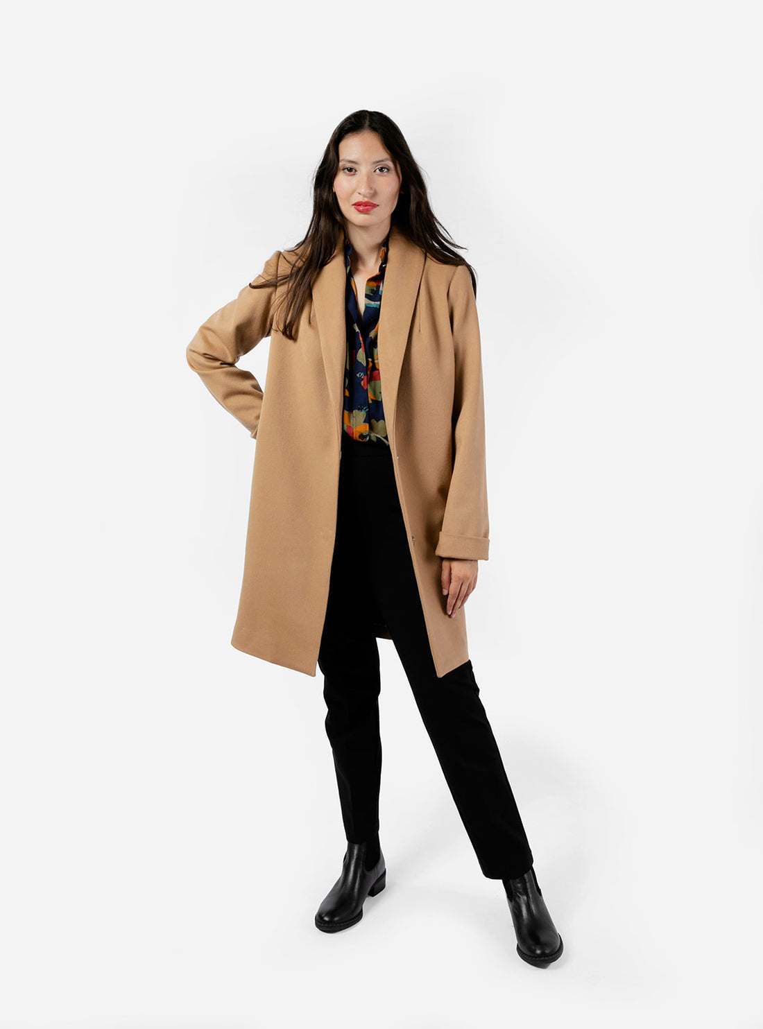 A stylish woman models a straight-cut knee-length women's dress coat in a rich camel hue, crafted from 100% wool for warmth and elegance. The classic camel coat features a timeless silhouette, perfectly complementing her black trousers and a patterned shirt. The combination creates a chic, sophisticated look that is both polished and comfortable, ideal for transitional weather. The soft texture of the wool adds an extra touch of luxury to her ensemble.