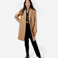 A stylish woman models a straight-cut knee-length women's dress coat in a rich camel hue, crafted from 100% wool for warmth and elegance. The classic camel coat features a timeless silhouette, perfectly complementing her black trousers and a patterned shirt. The combination creates a chic, sophisticated look that is both polished and comfortable, ideal for transitional weather. The soft texture of the wool adds an extra touch of luxury to her ensemble.