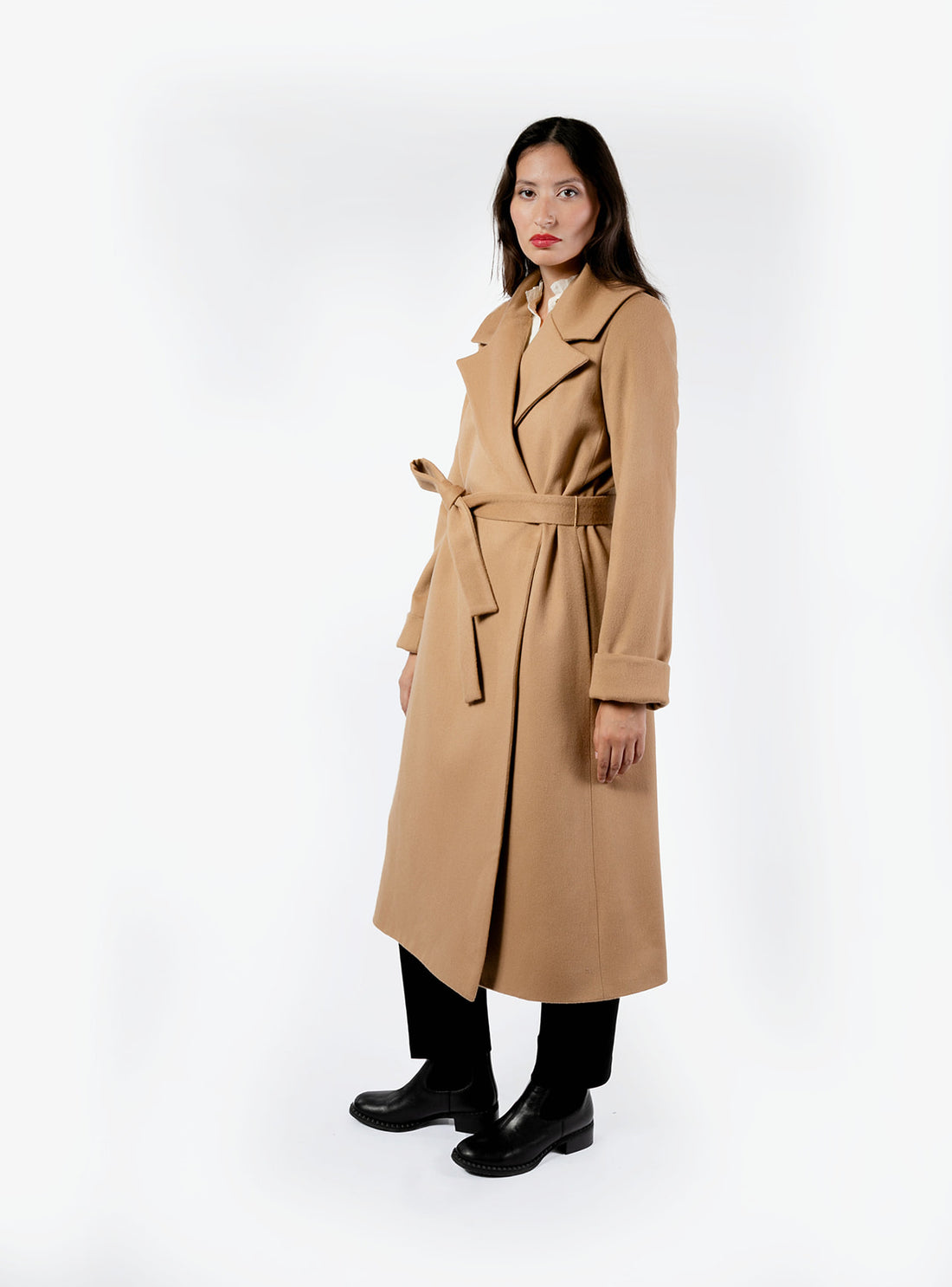 Three quarter view of a straight-cut, full-length women's belted coat in a camel color, made from 100% wool. The long camel coat features an oversized notched lapel, long cuffed sleeves, and is paired with sleek black trousers and black boots.