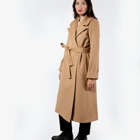Three quarter view of a straight-cut, full-length women's belted coat in a camel color, made from 100% wool. The long camel coat features an oversized notched lapel, long cuffed sleeves, and is paired with sleek black trousers and black boots.