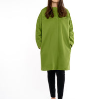 A woman confidently models a collarless, knee-length green wool coat made from high-quality RWS wool. The coat showcases clean lines and a streamlined silhouette, reflecting the bold, youthful spirit of the 1960s and striking green color. Its minimalist design and contemporary twist highlight a chic and timeless style, perfect for modern fashion.