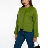A woman stands in 3/4 view, wearing a modern-cut green lady jacket with drop patch pockets paired with stylish jeans. She holds a vintage black purse, adding a touch of classic elegance to her contemporary outfit. The scene captures her confident stance against a neutral background.
