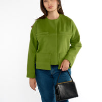 A woman stands wearing a modern-cut green lady jacket with drop patch pockets paired with stylish jeans. She holds a vintage black purse, and bold gold earrings, adding a touch of classic elegance to her contemporary green lady jacket. The scene captures her confident stance against a white background.