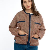 A woman stands confidently with her hands in the pockets of a modern-styled tweed lady jacket, trimmed with navy grosgrain. The tweed lady jacket features classic styling with modern drop patch pockets, adding a stylish touch to her classic outfit paired with jeans.