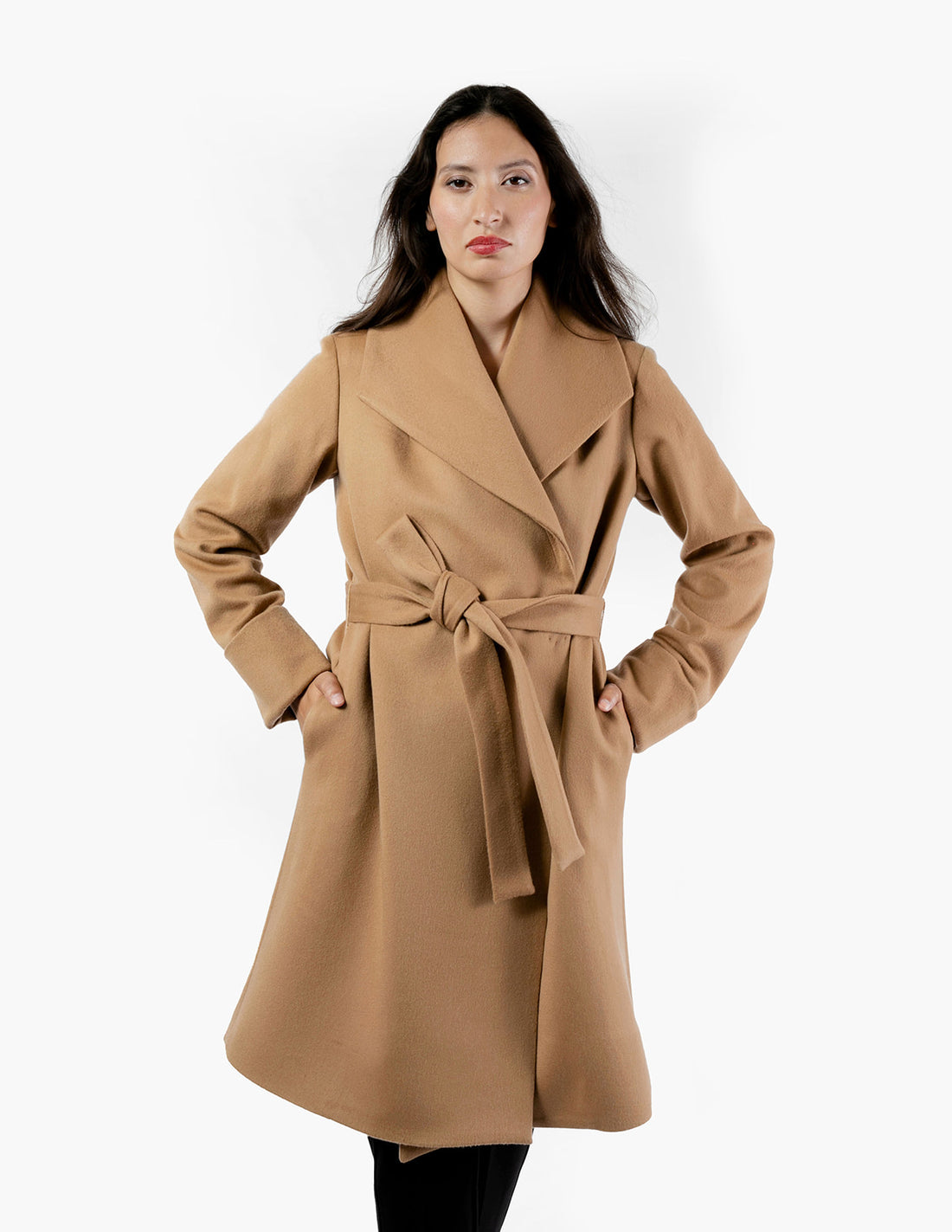 A woman wearing a knee-length wool camel coat with an oversized collar and a belted waist. The camel coat features cuffed sleeves, and the women's hands are tucked into the pockets. She stands confidently, showcasing the coat's elegant silhouette, beautiful wool fabric and camel color.