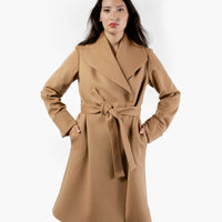 A woman wearing a knee-length wool camel coat with an oversized collar and a belted waist. The camel coat features cuffed sleeves, and the women's hands are tucked into the pockets. She stands confidently, showcasing the coat's elegant silhouette, beautiful wool fabric and camel color.