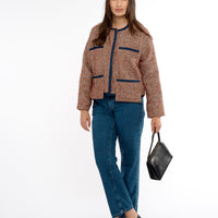A woman stands poised in a tweed lady jacket trimmed with navy grosgrain, paired with jeans. She carries a vintage black bag, and the jacket features stylish drop pockets, adding to her chic, casual look against a neutral background.