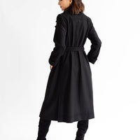 Full length photo of the back view pf a long black wool coat on a model. Photo shows calf length hem, belted waist, long, cuffed sleeves, and back walking vent.