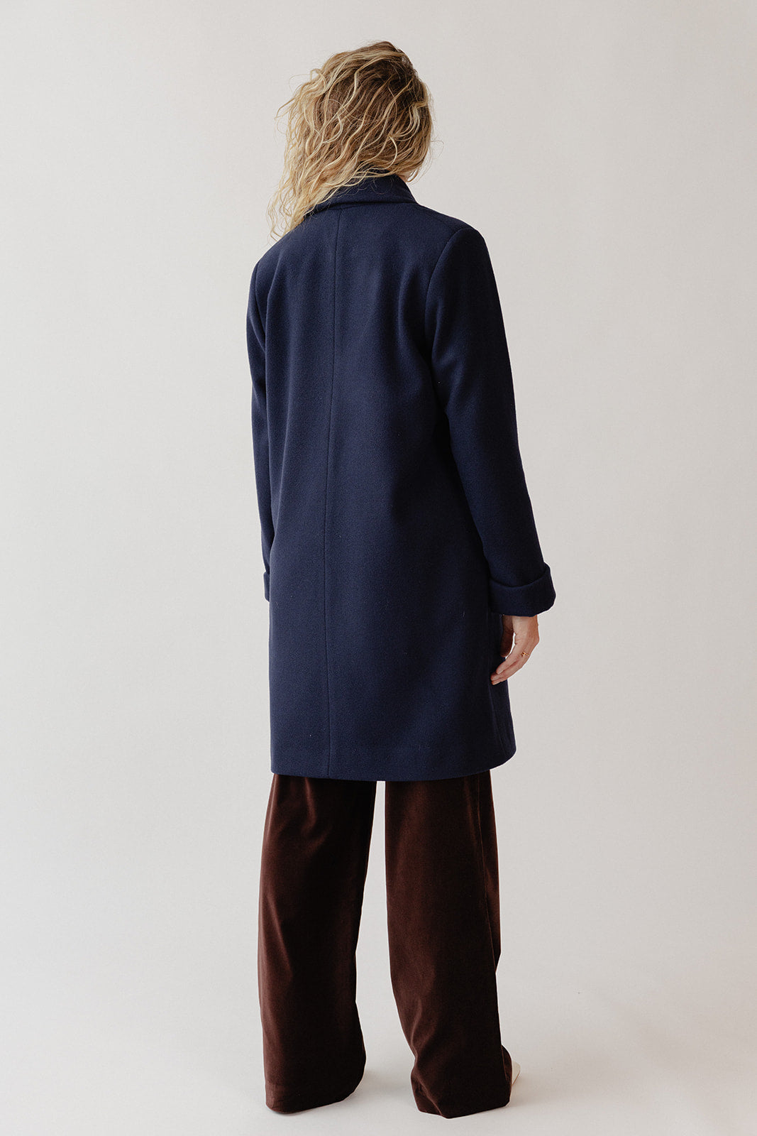 Back view of a woman standing wearing a navy blue wool coat and brown velvet pants.