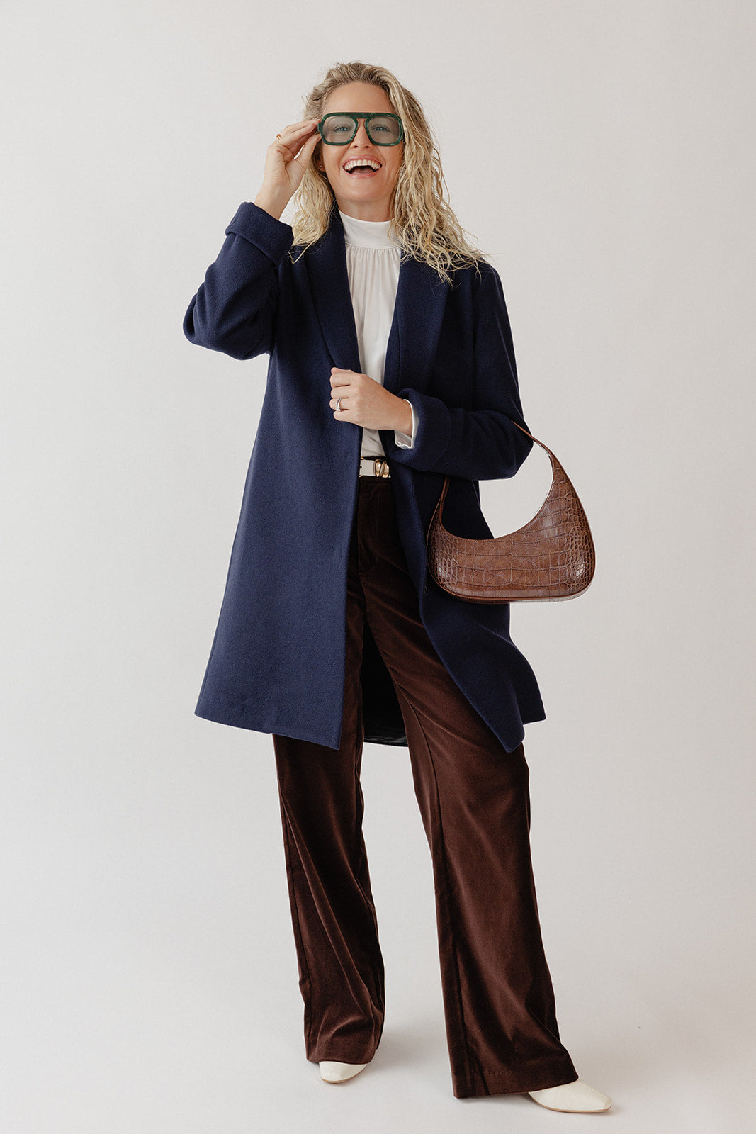 Woman laughing wearing a Navy blue wool coat, white blouse, brown velvet pants, and fun green eyeglasses