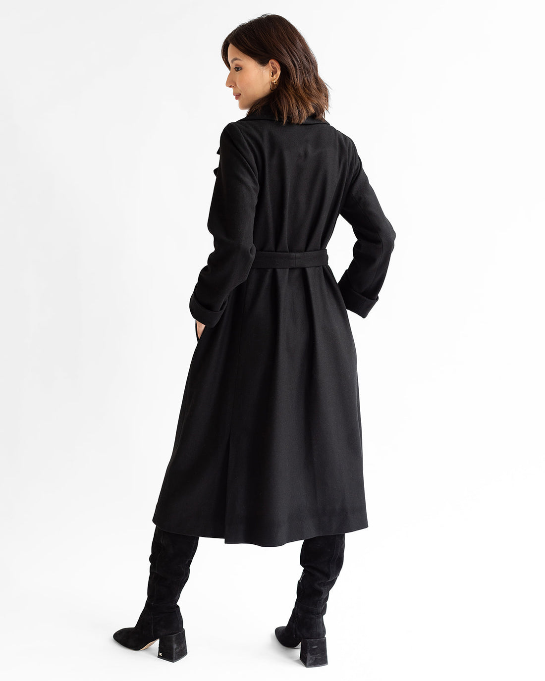Full length photo of the back view pf a long black wool coat on a model. Photo shows calf length hem, belted waist, long, cuffed sleeves, and back walking vent.